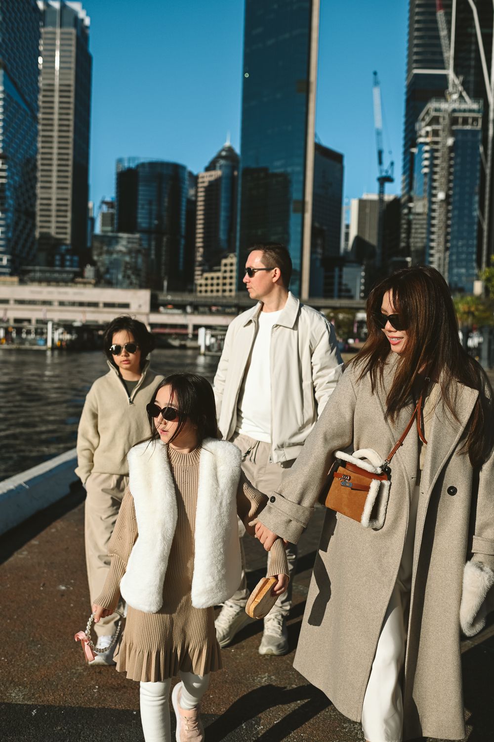 A stylish family in matching beige and white coats walks along a waterfront with city skyscrapers in the background.