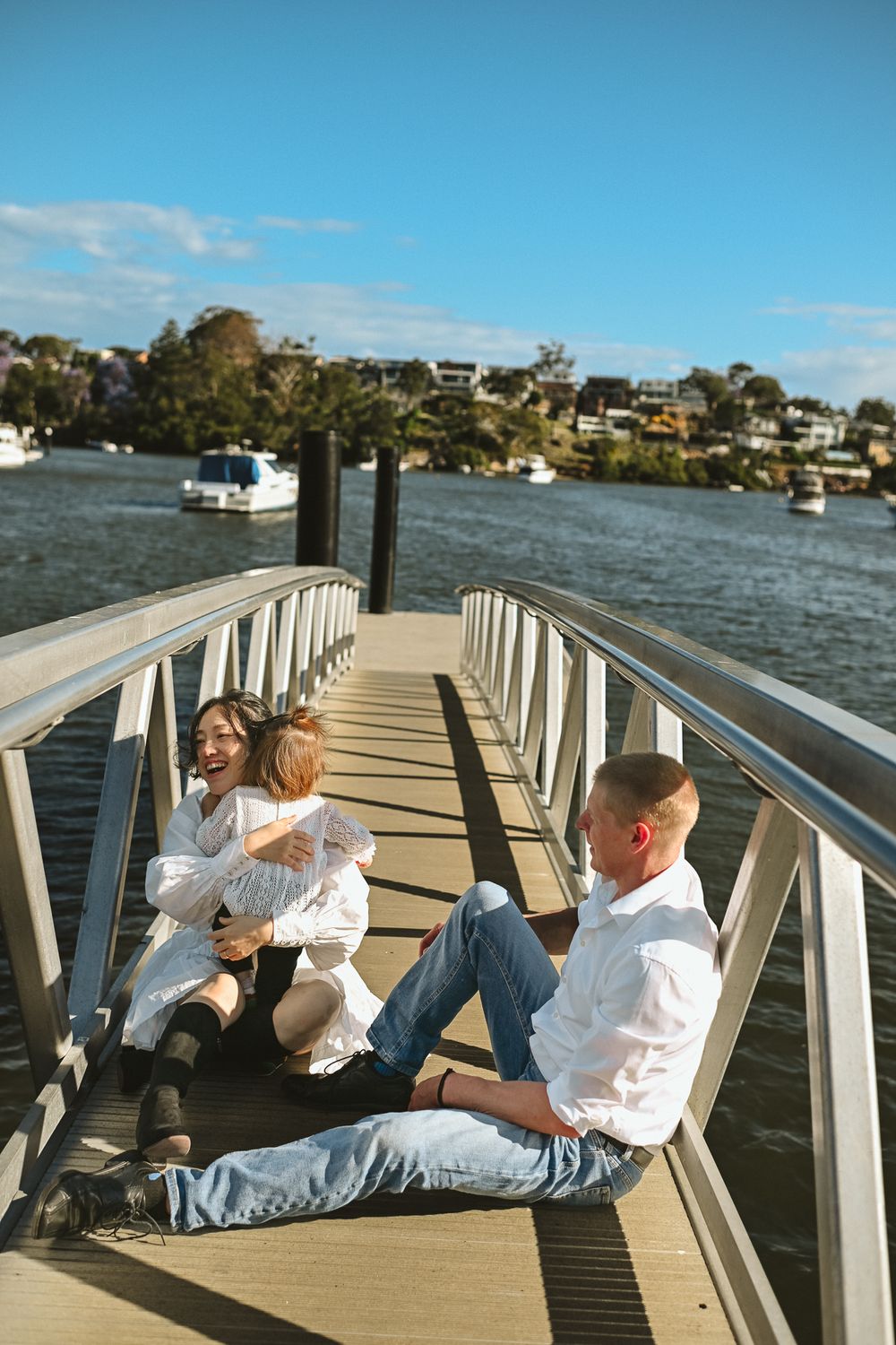 Couple in white shirts relaxing together on a wooden dock overlooking a scenic waterfront harbor on a sunny day.