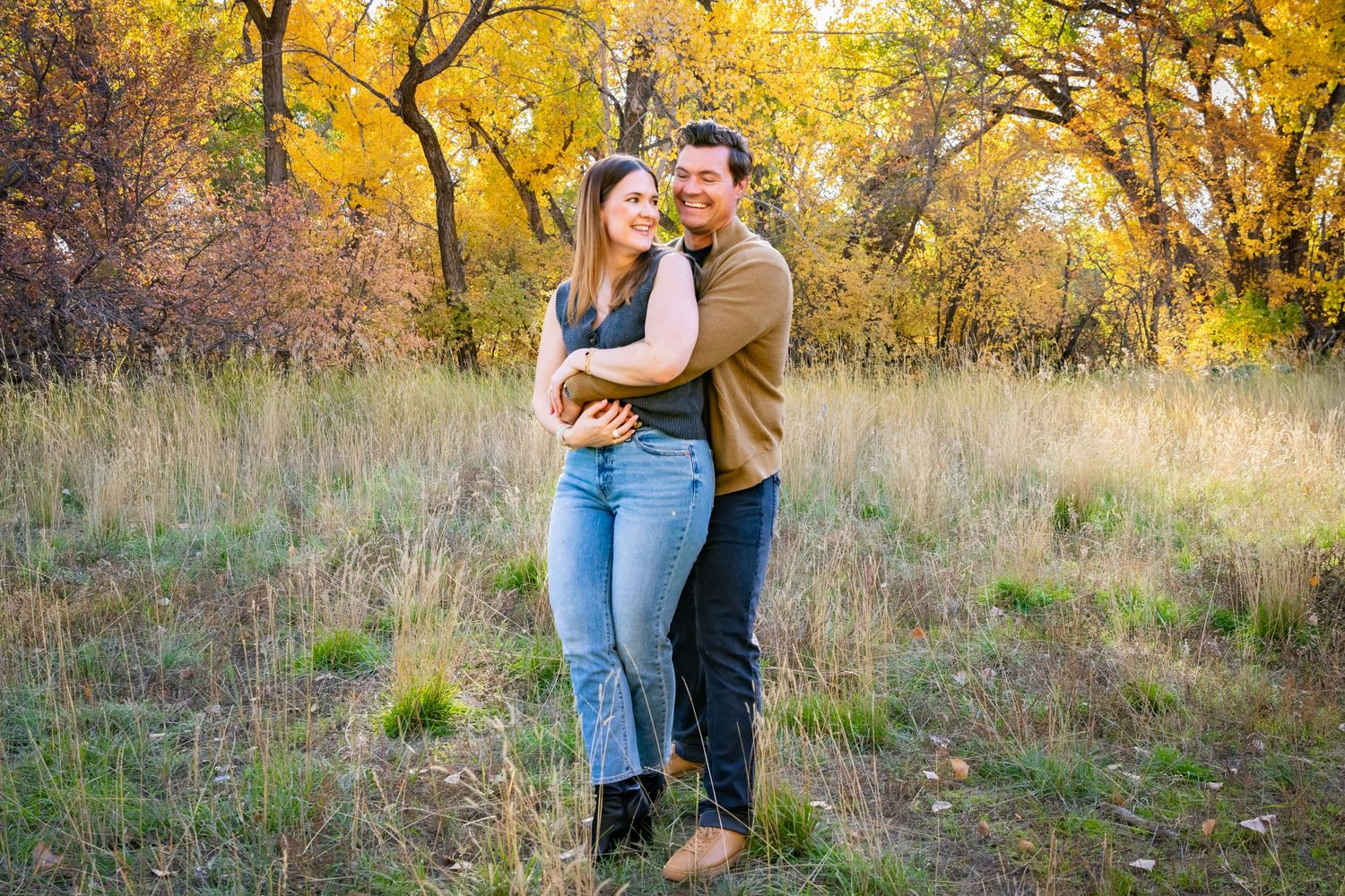 a couple holding each other in a field with autumn trees in the background.