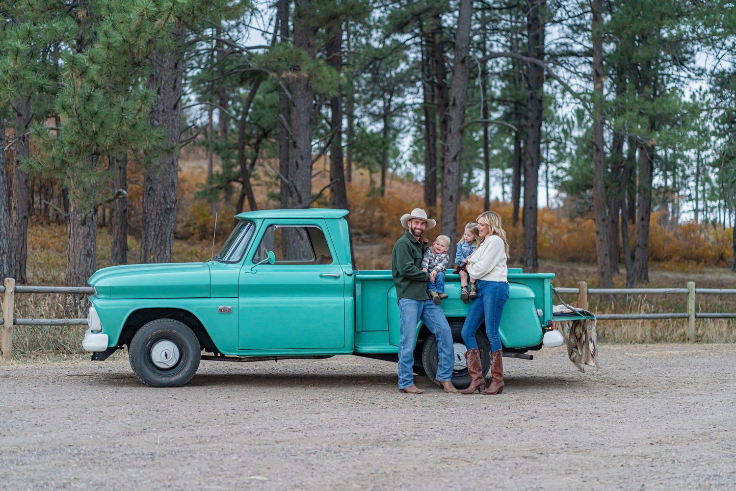 Couple stands together next to a vintage turquoise pickup truck in a pine forest setting.