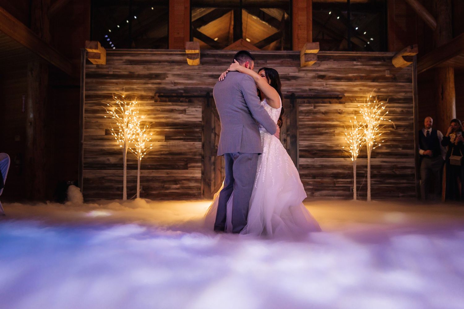 Couple shares their first dance in a rustic barn venue with romantic lighting and fog effects.