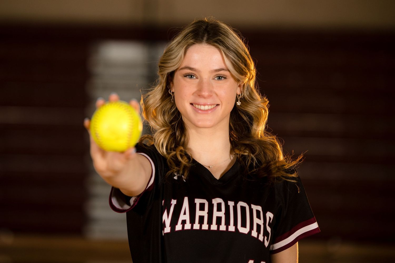 A softball player in Warriors uniform holding up a yellow softball with a bright smile.