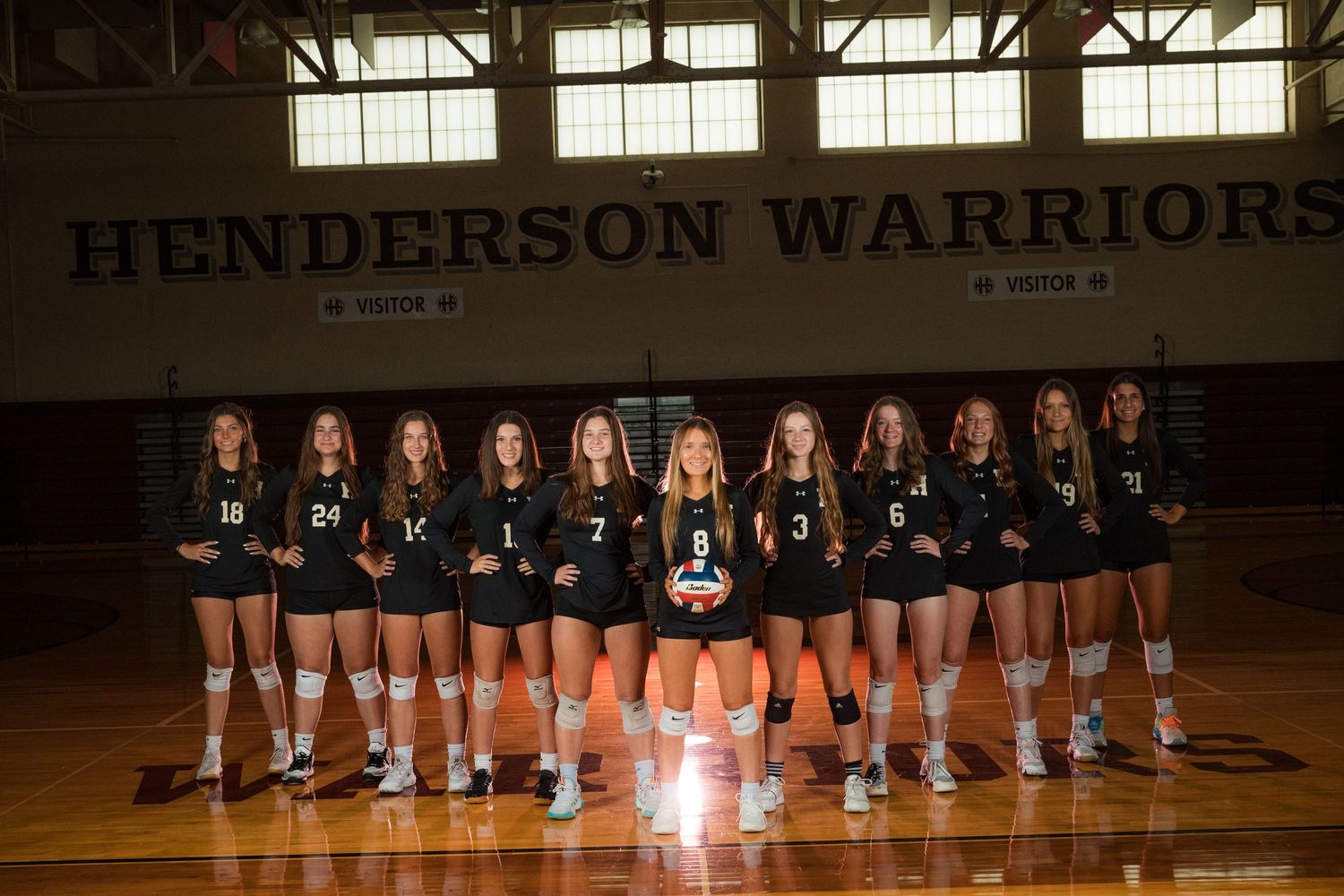 High school volleyball team poses in formation on gymnasium court with Henderson Warriors banner.