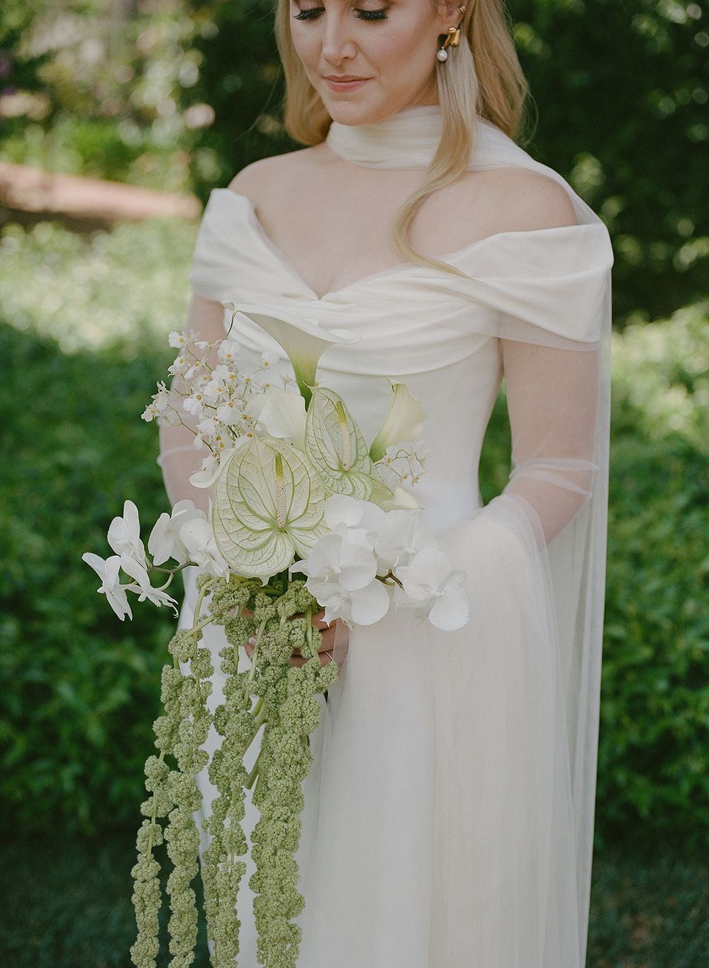 Elegant off-shoulder white wedding gown with cascading greenery and white floral bouquet in an outdoor garden setting.