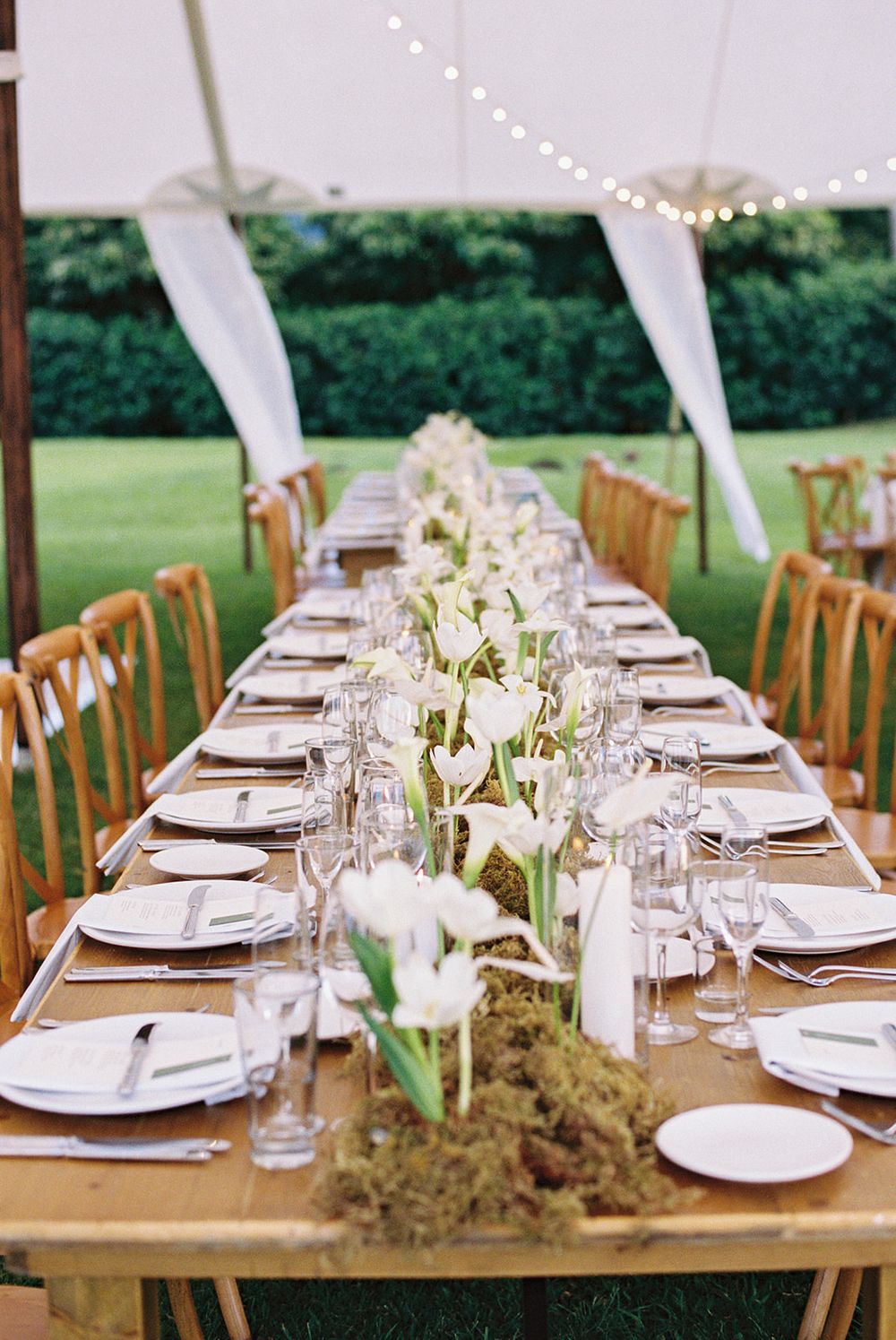 Elegant outdoor wedding table setting with white floral centerpieces and rustic wooden chairs under string lights.