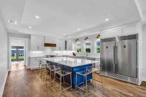 Modern white kitchen with large island, blue bar stools, and hardwood floors illuminated by recessed lighting.