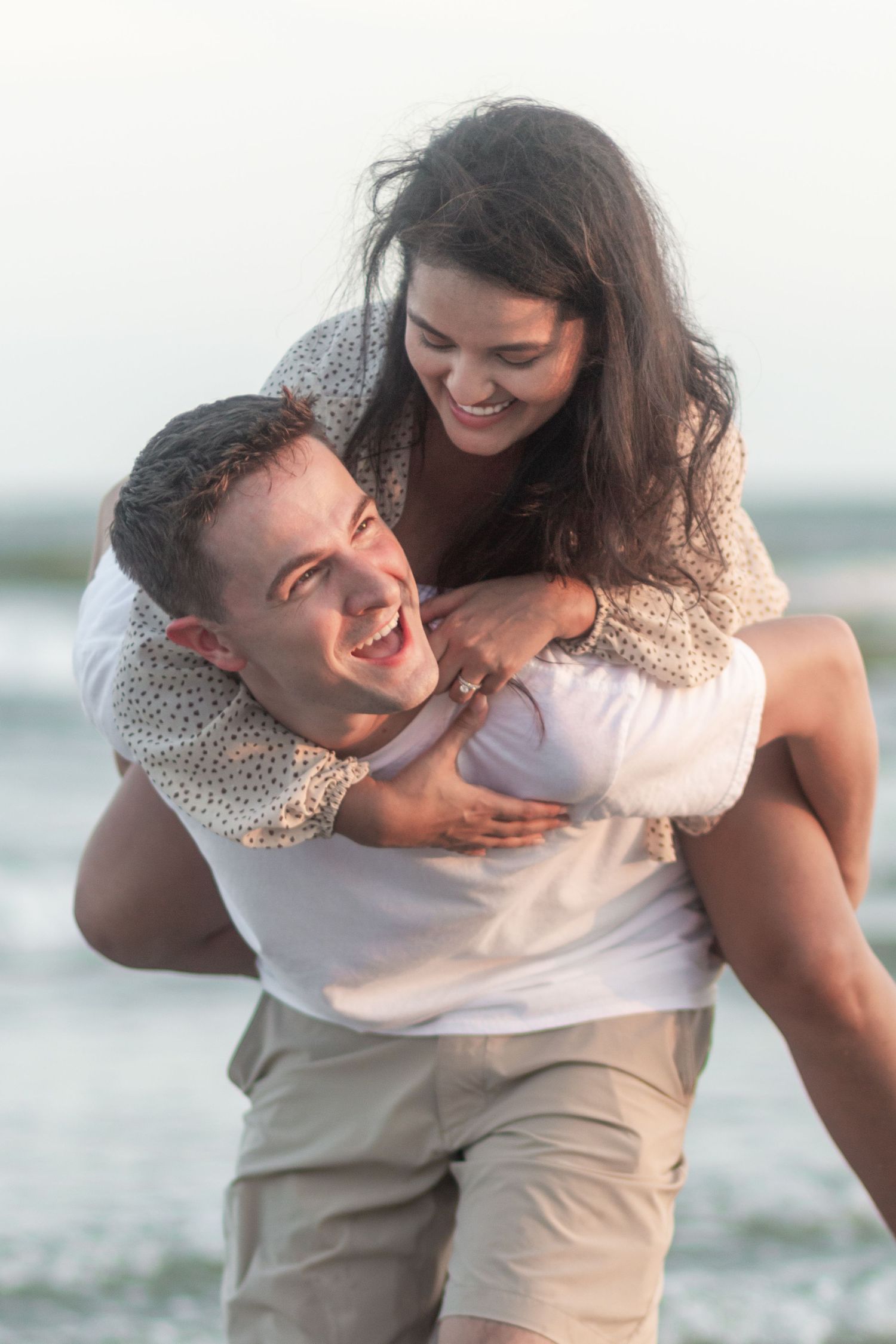 Happy couple enjoying a piggyback ride on the beach with ocean waves behind them.