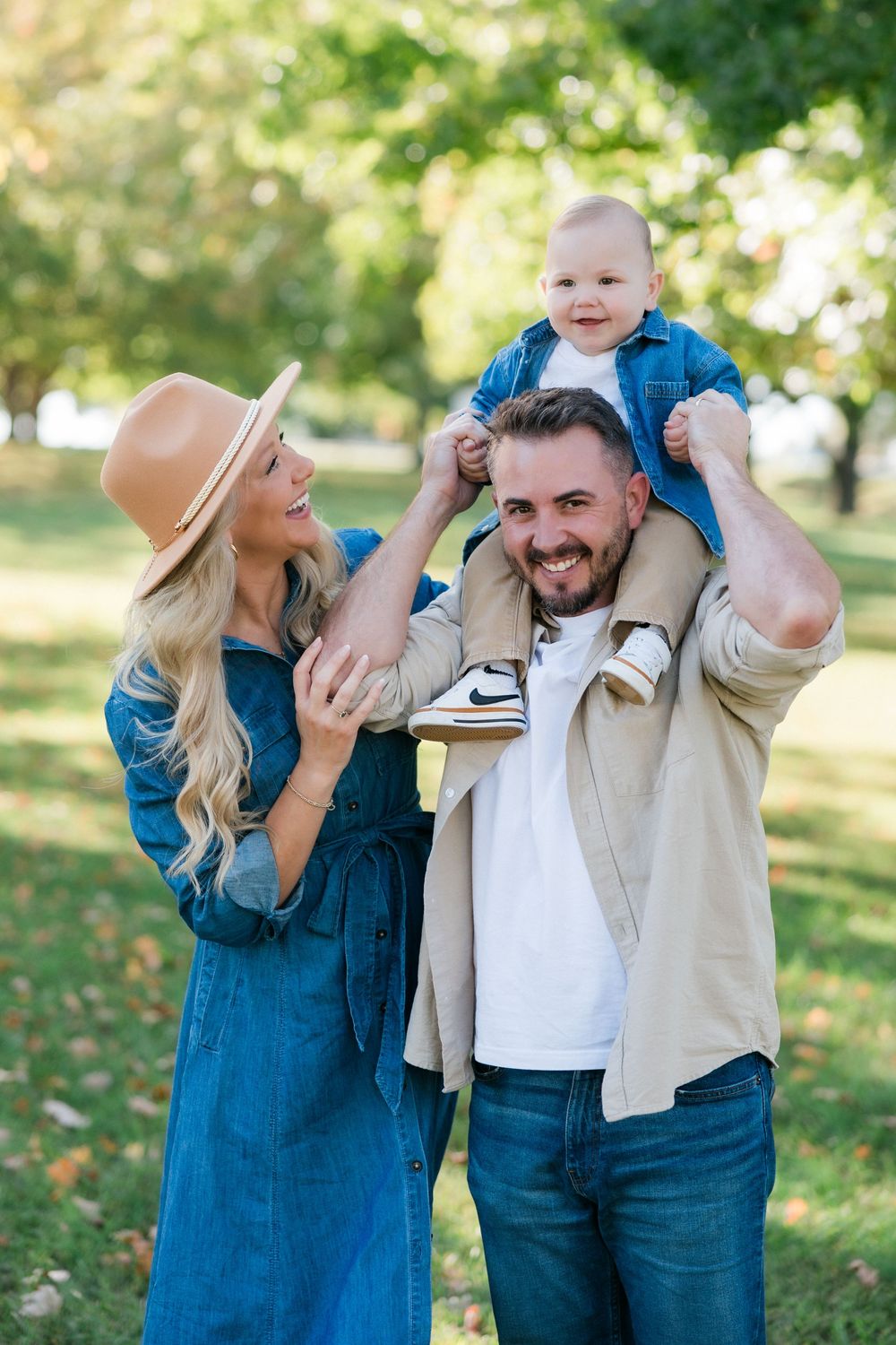 Family portrait in autumn park with child sitting on shoulders and mom wearing blue dress and brown hat.