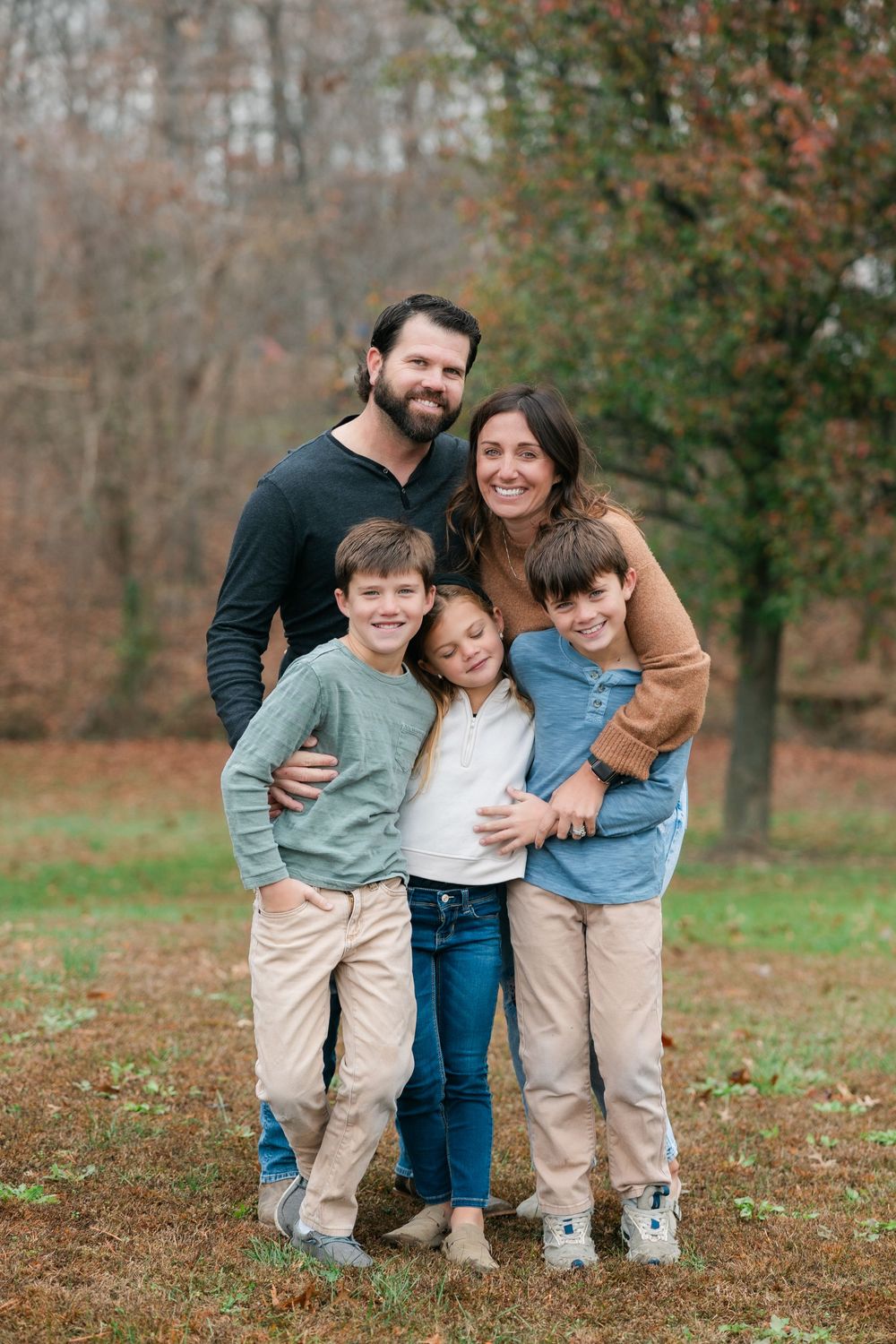 A smiling family of five poses together outdoors on an autumn day with fall foliage in the background.