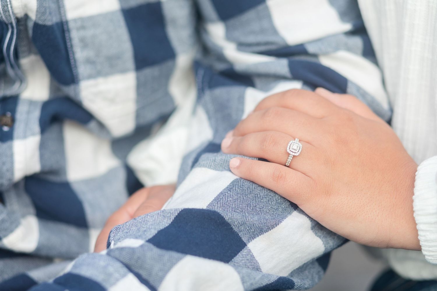 Close up of diamond engagement ring on hand against blue and white plaid fabric background.