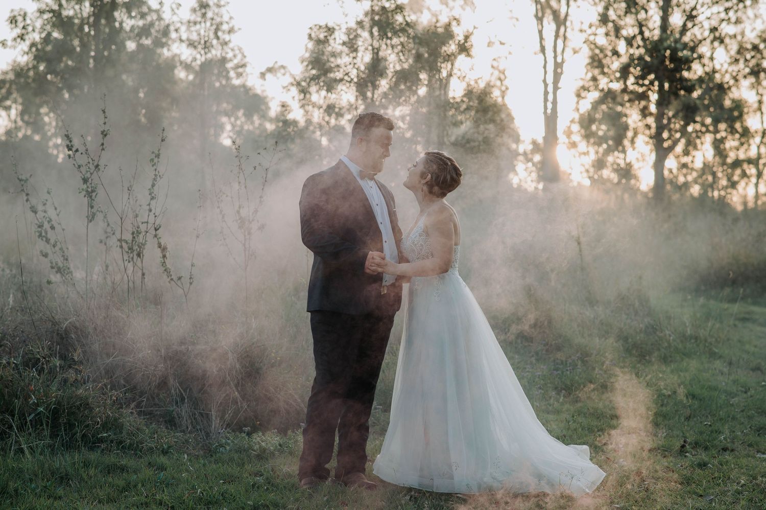 A romantic wedding couple embraces in a misty field at sunset with ethereal golden light surrounding them.
