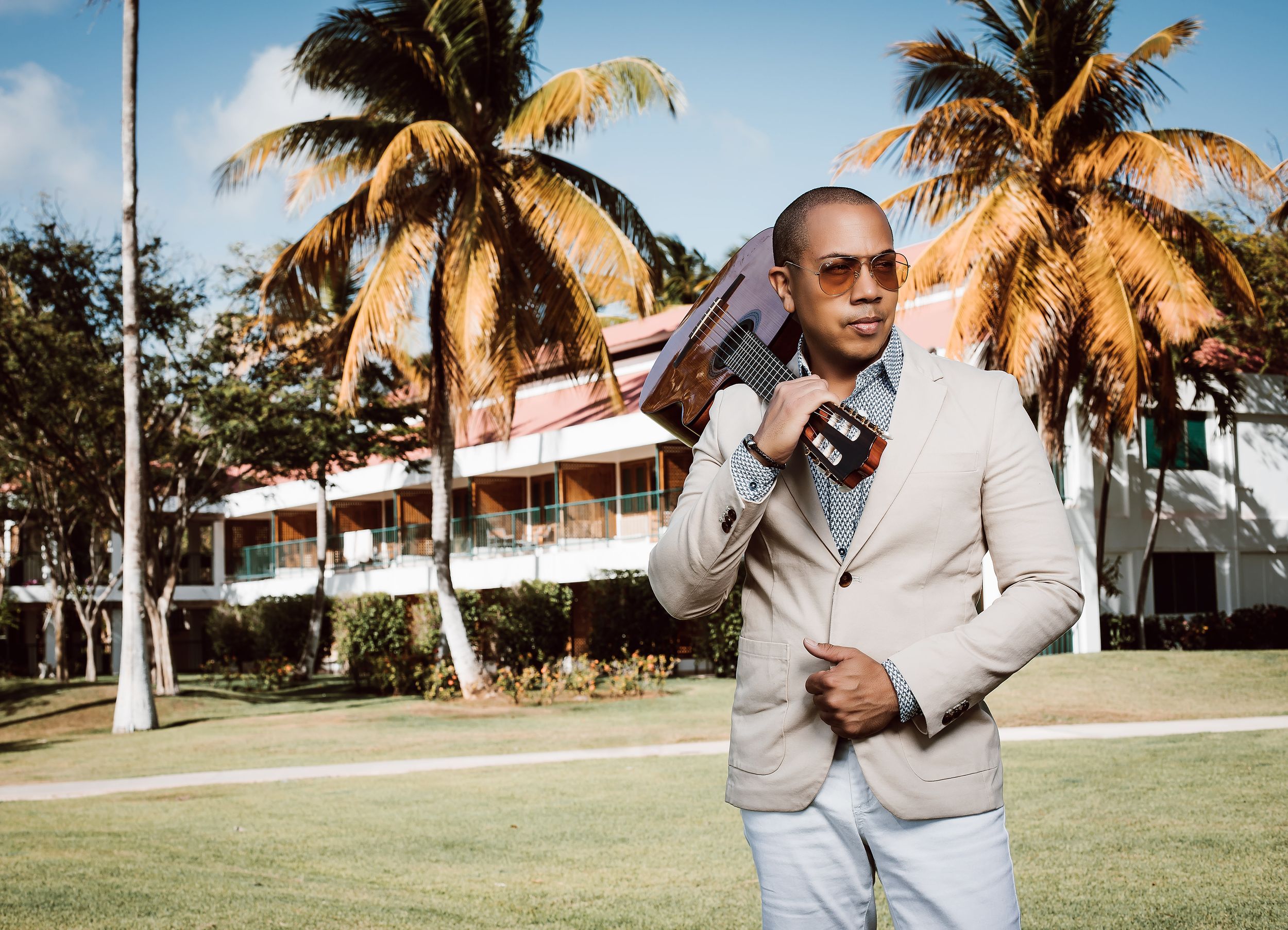 A person in a cream colored suit poses casually in front of palm trees and white buildings on a sunny day.