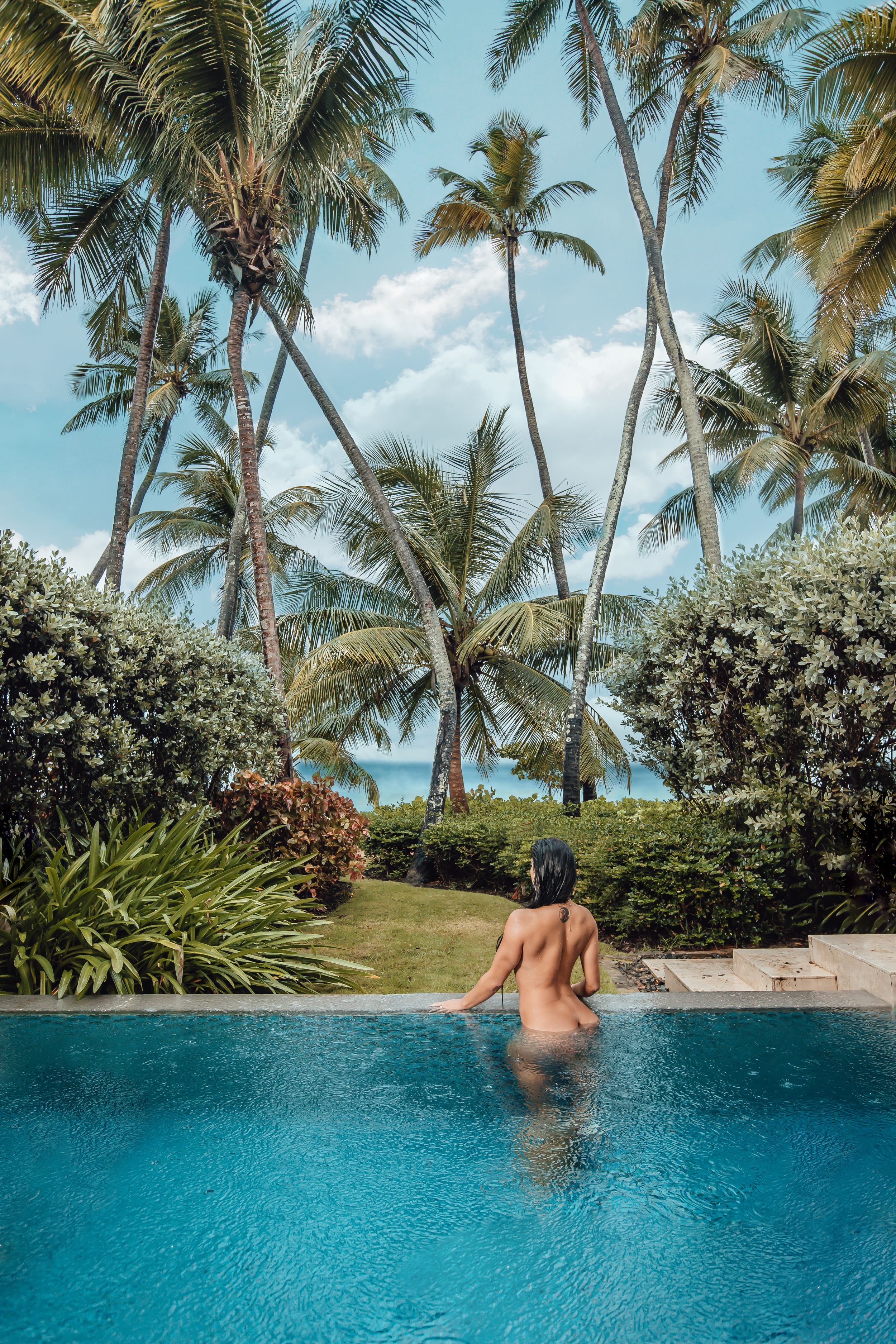 Person relaxing in infinity pool overlooking tropical palm trees and ocean view.