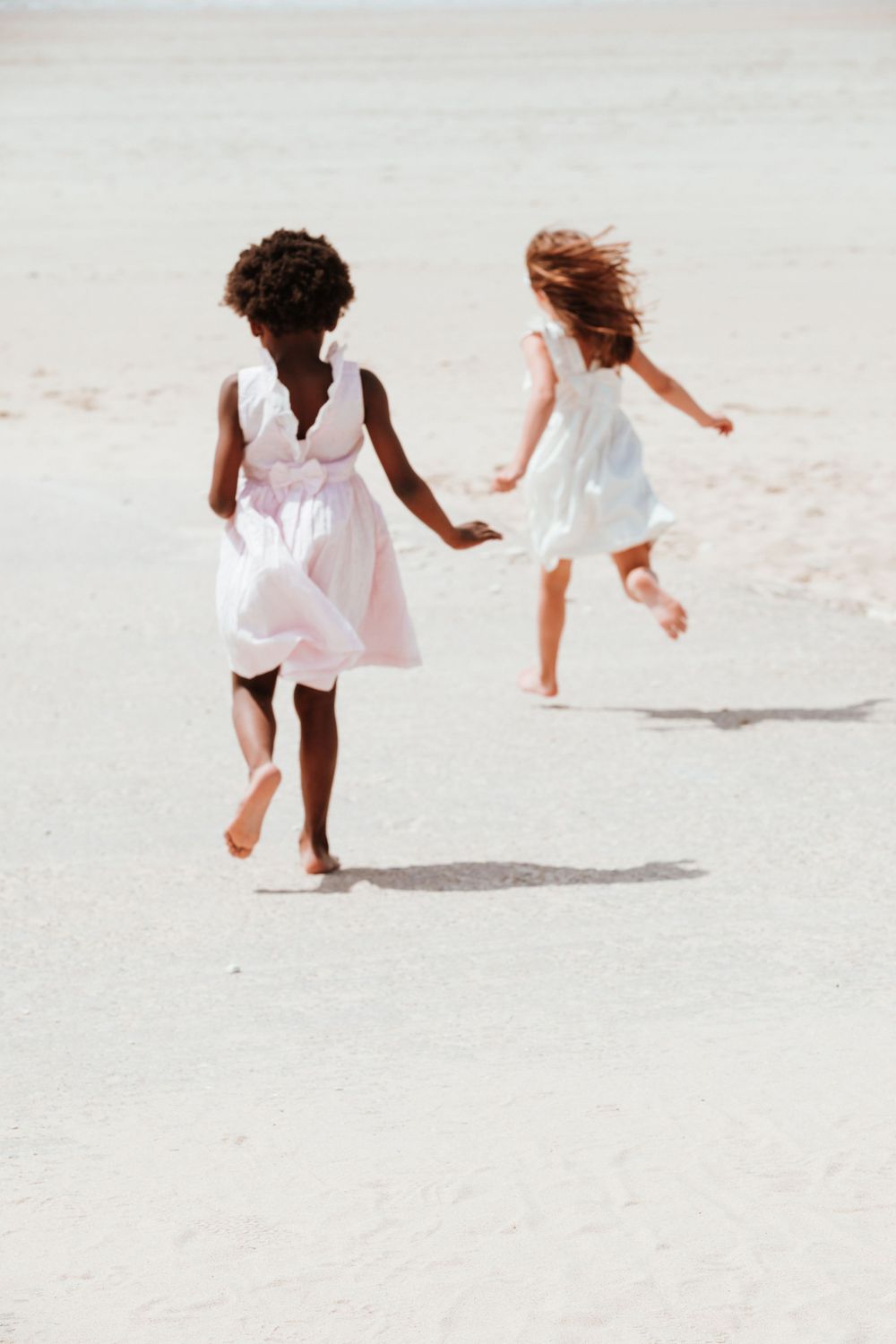 Two figures in white dresses running together on sandy beach.