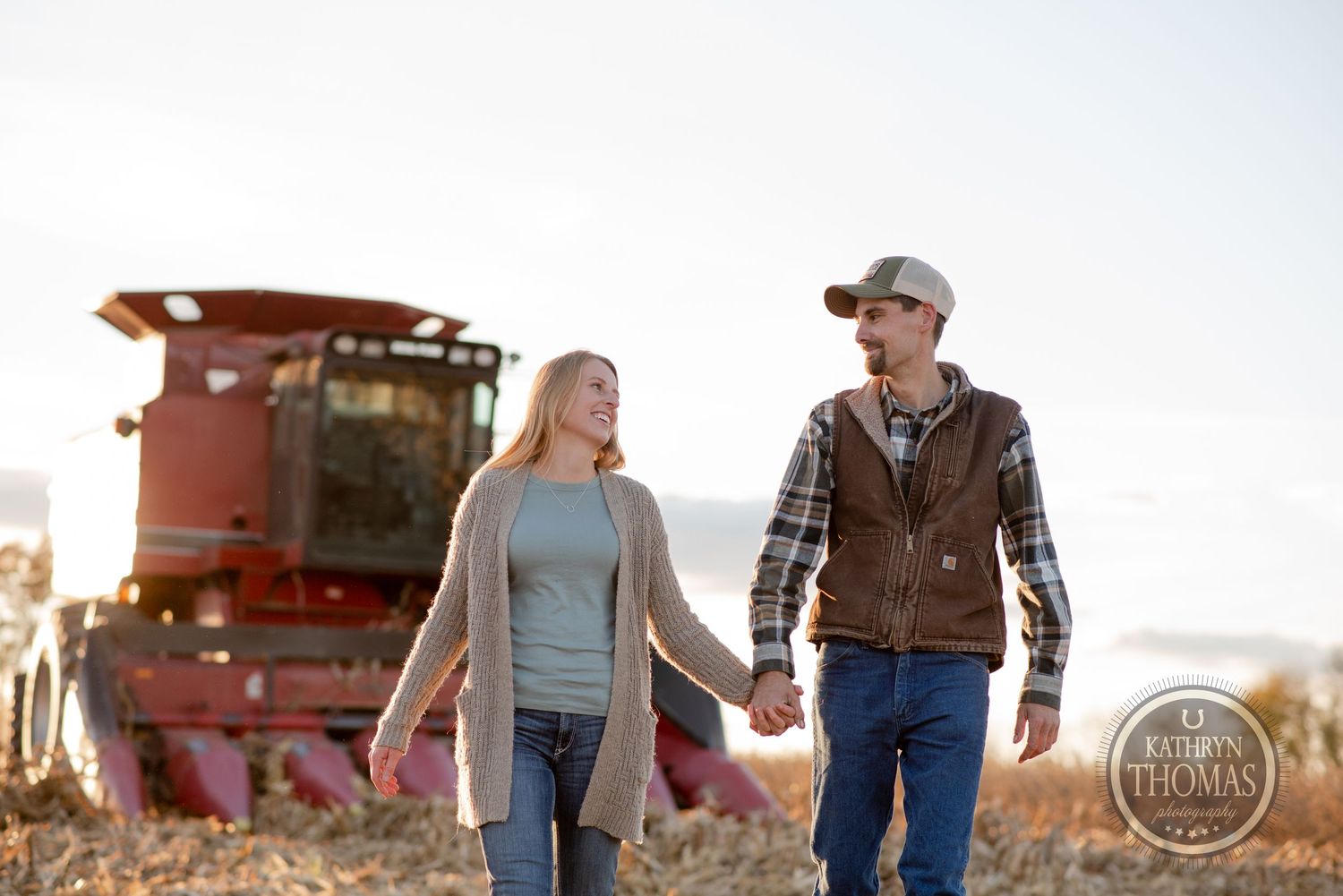 Couple walking hand in hand through a harvested field with red combine harvester in background.