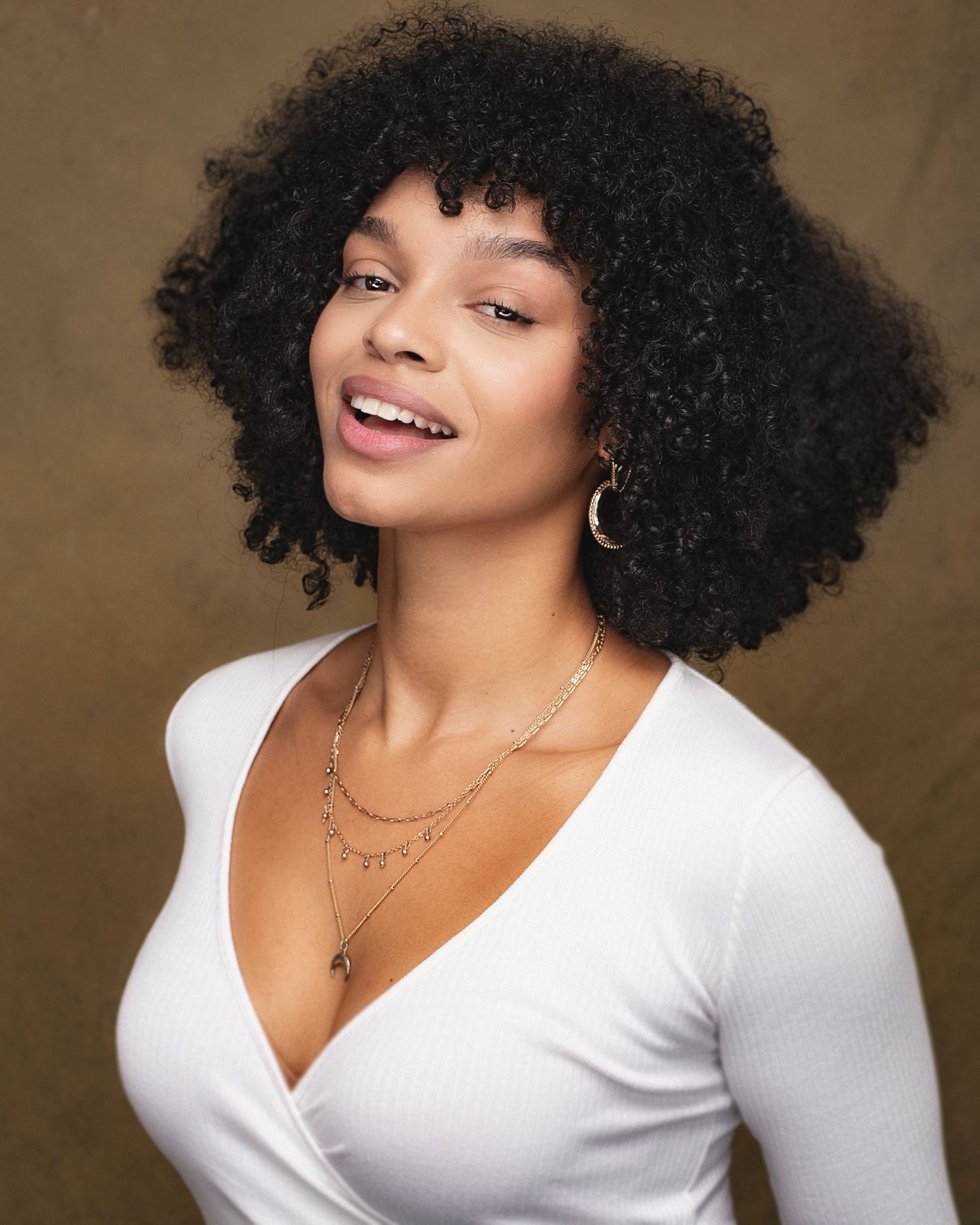 Studio portrait showing someone in a white top with natural curly hair and layered necklaces.