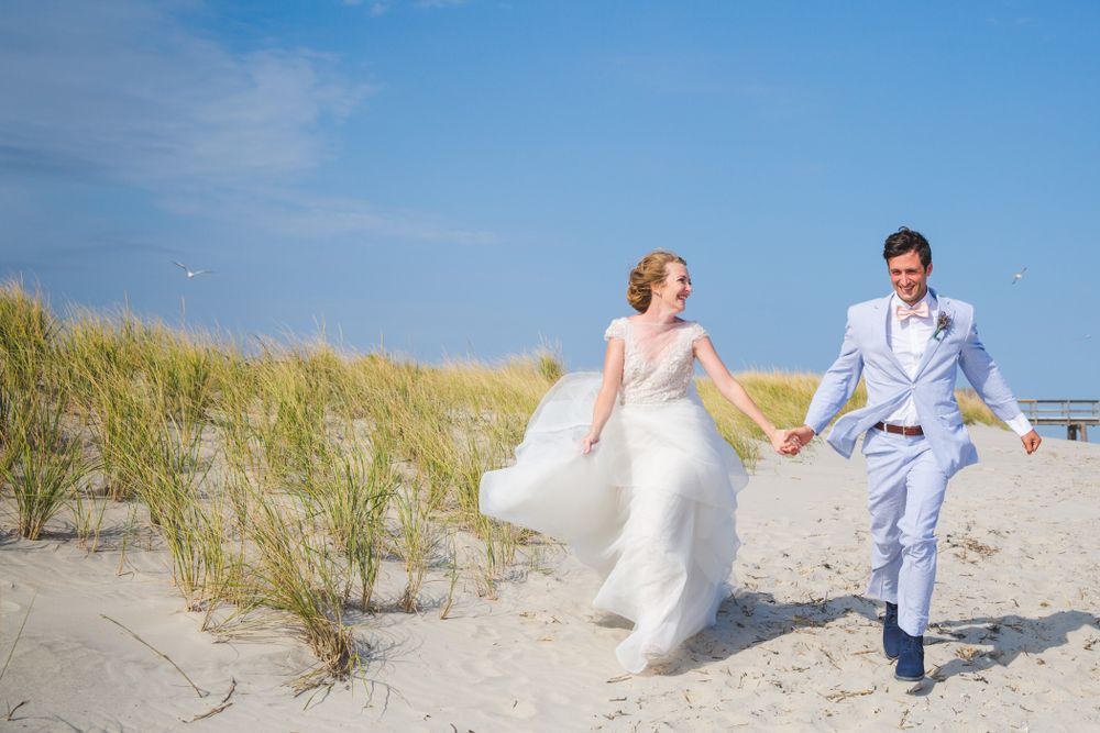 Couple runs joyfully along beach dunes in wedding attire.