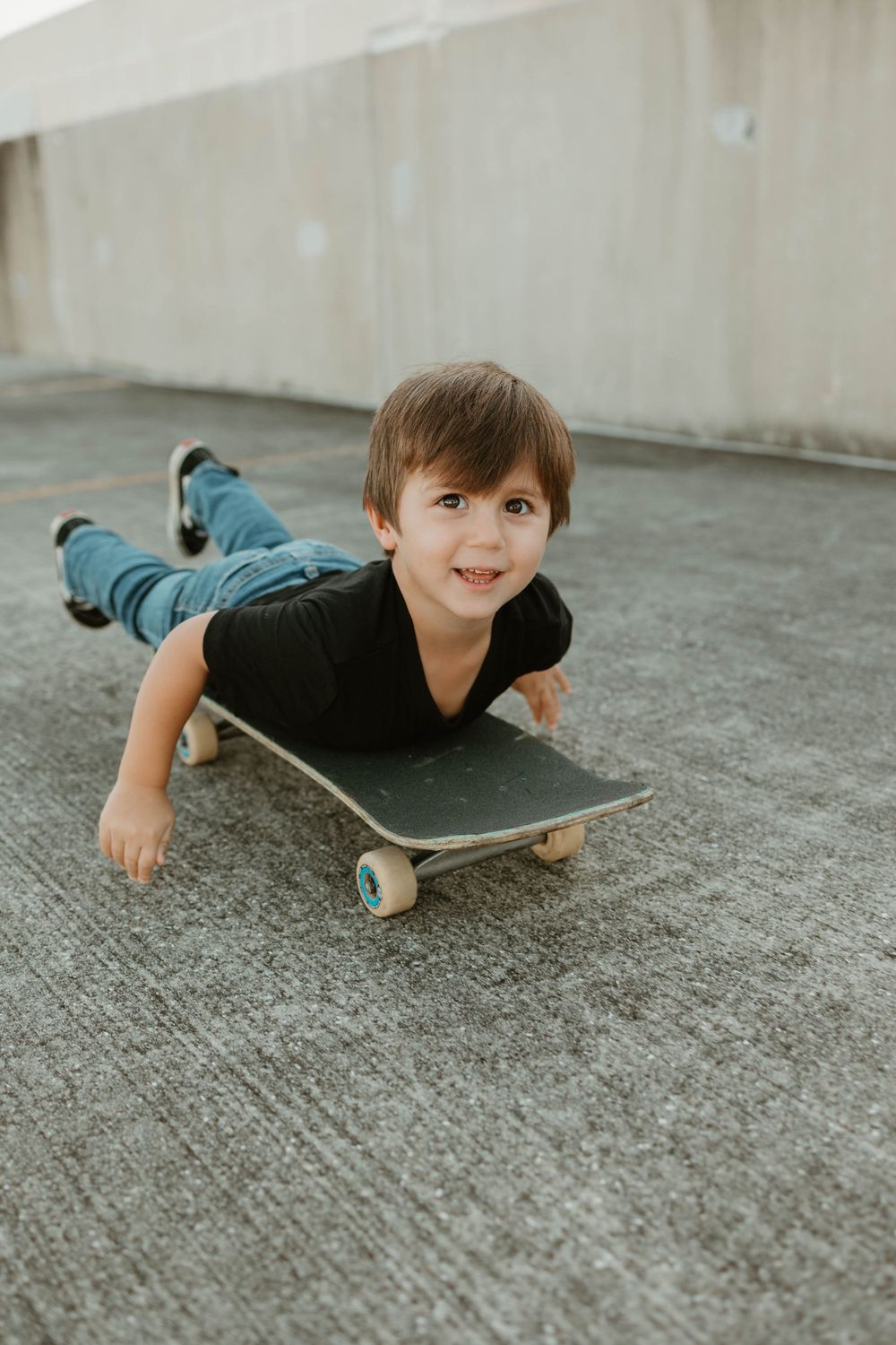 A young skateboarder lying flat on a skateboard on concrete ground, demonstrating a unique skating position.