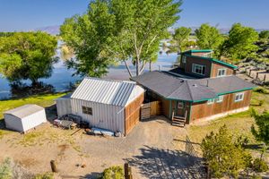 Aerial view of waterfront cabin with metal roof, large trees, and scenic lake backdrop.