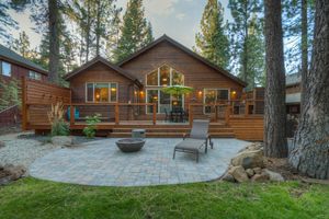 Rustic wood cabin exterior with large deck, patio seating, and tall pine trees.
