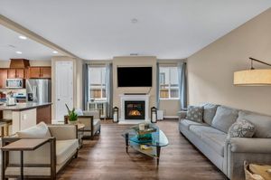 Spacious living room with gray sofa, fireplace, wall-mounted TV, and open view to modern kitchen.