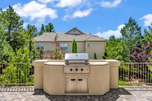 Built-in outdoor kitchen with stainless steel grill on a patio surrounded by lush trees and an iron fence.