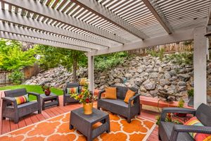 Covered patio with white pergola, wicker furniture, colorful rug, and decorative rock wall backdrop.