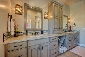 Double vanity bathroom with gray cabinetry, granite countertops, dual mirrors, decorative sconces, and built-in storage tower.