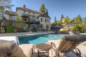 Luxury outdoor pool area with lounge chairs, patio umbrella, and large multi-story home surrounded by tall trees.