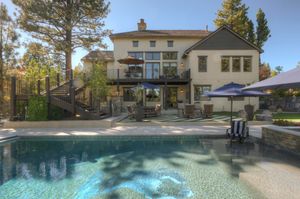 Resort-style backyard pool with stone patio, outdoor seating, umbrella, and elegant two-story home with balcony.
