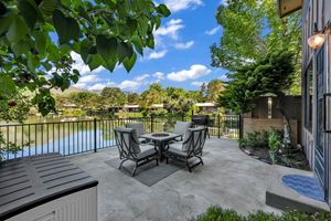 Outdoor patio with dining set overlooking a tranquil lake surrounded by lush greenery under a blue sky.