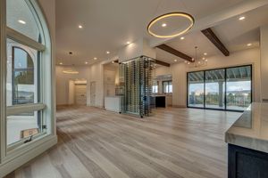 Spacious open-concept living area with light hardwood floors, exposed beams, wine display, and large windows.