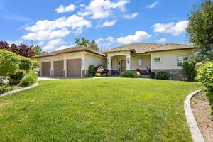 Ranch-style home with three-car garage, large green lawn, mature trees, and stone accents under sunny blue sky.