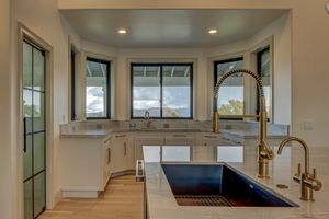 Bright kitchen corner with white cabinetry, marble countertops, large black undermount sink, and gold industrial-style faucet.