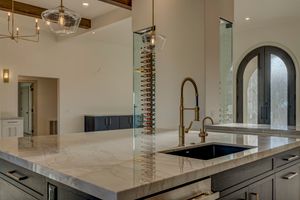 Close-up of marble kitchen island with black undermount sink, gold faucet, pendant lights, and glass wine rack in background.