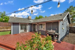 Red wood deck with BBQ grill attached to a gray single-story home with string lights and lush green backyard.