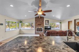 Open-concept living room with vaulted ceiling, stone fireplace, ceiling fan, and dark granite kitchen counter in foreground.