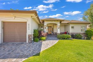 Elegant single-story stucco home with arched entry, paver driveway, stone accents, and lush green front lawn.