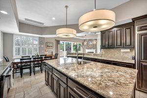 Spacious kitchen with dark cabinetry, large granite island, drum pendant lights, and a dining area beyond.