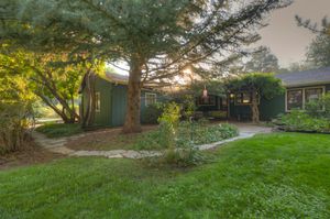 Green ranch-style home surrounded by mature trees with a winding garden path at sunset.