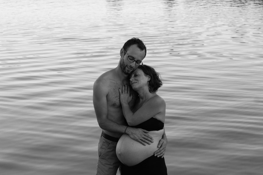 An intimate black and white photograph captures a loving embrace between expectant parents near water.
