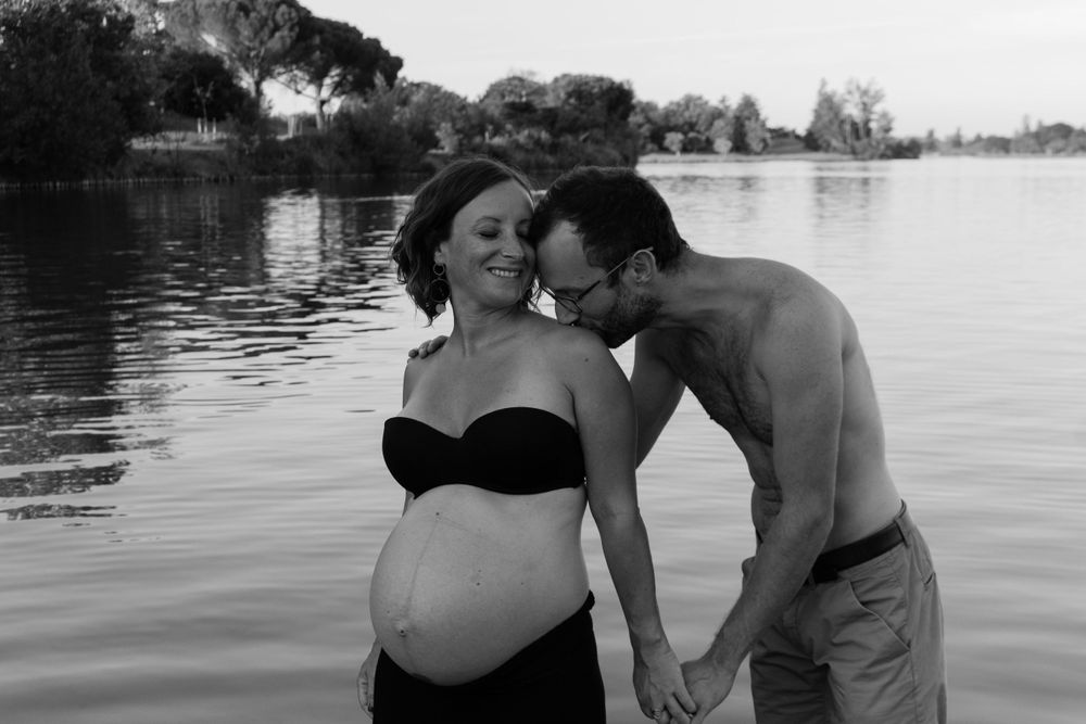 A romantic maternity photoshoot by the lake shows a tender moment between an expectant couple.
