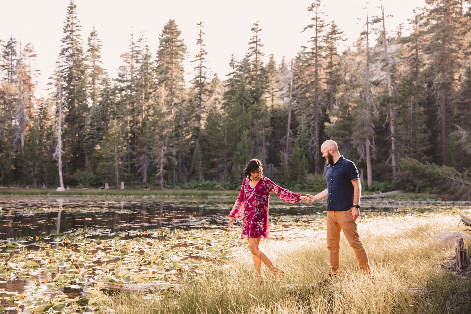 Couple holding hands walking along a scenic lakeside path surrounded by tall pine trees.