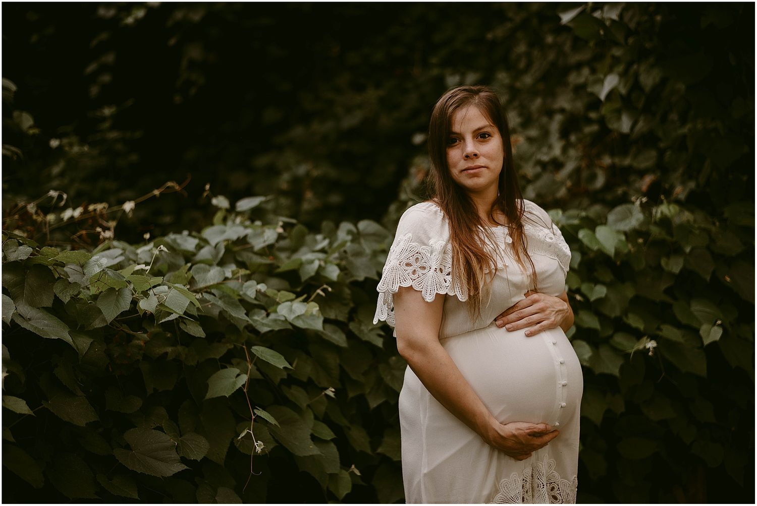 Maternity photo in white lace dress against leafy garden backdrop during golden hour.
