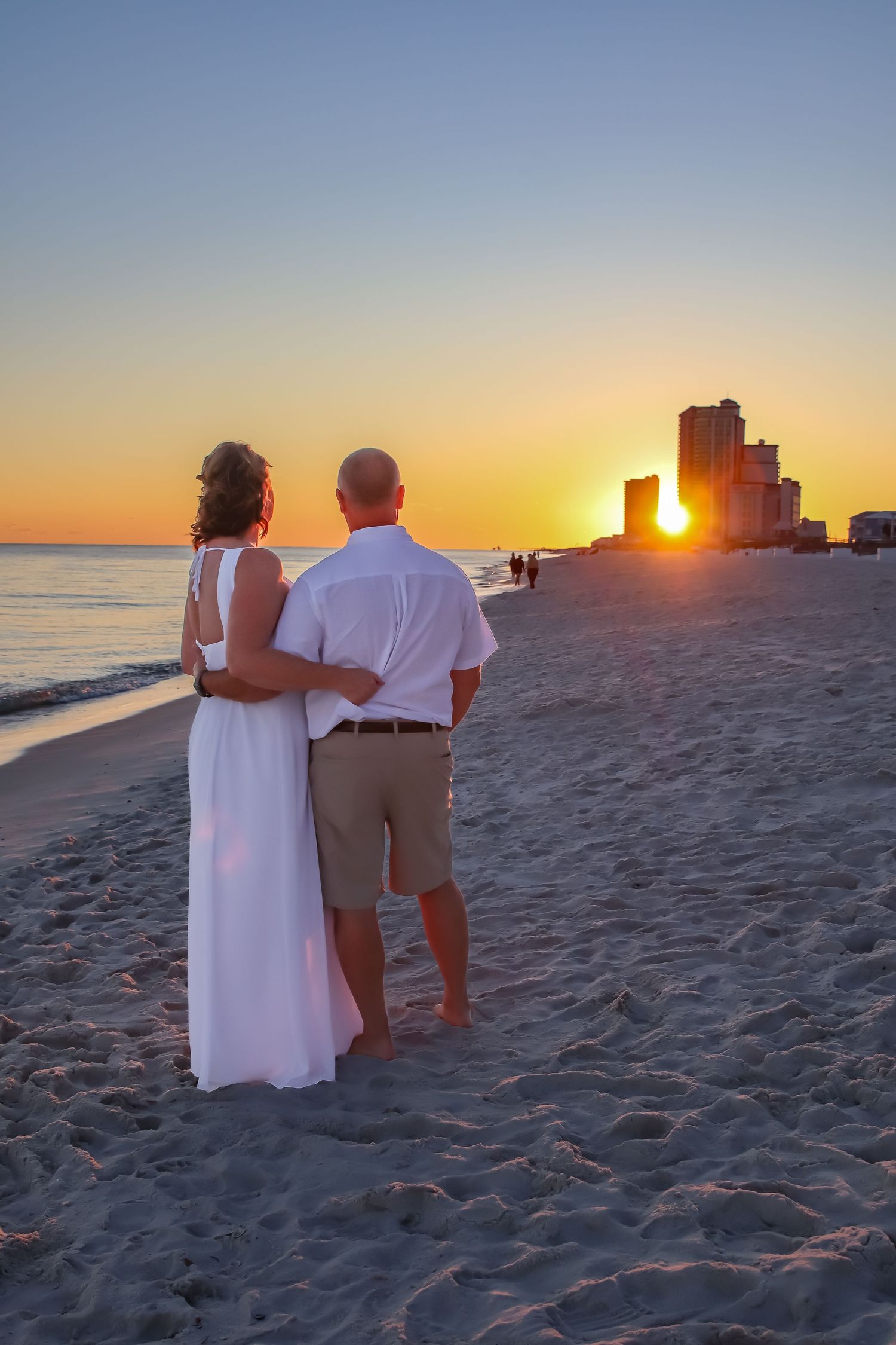 A couple watches the sunset on a beach with high-rise buildings silhouetted against the orange sky.