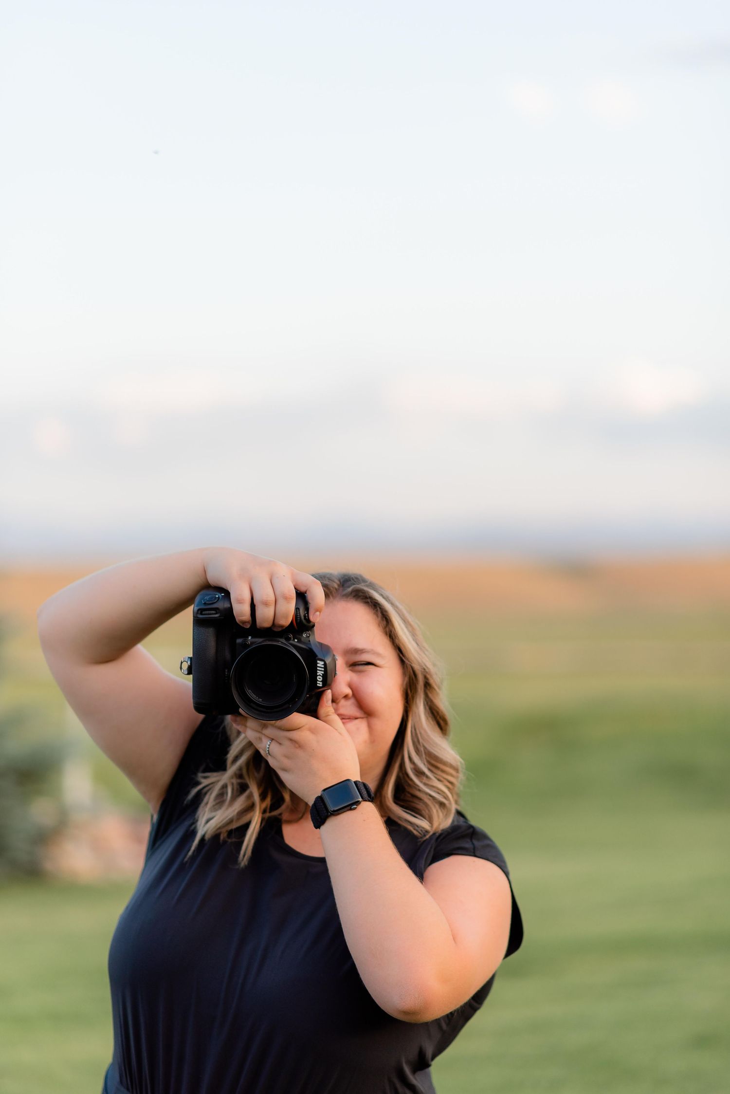 Woman photographer smiling while holding a camera outdoors in a green field.
