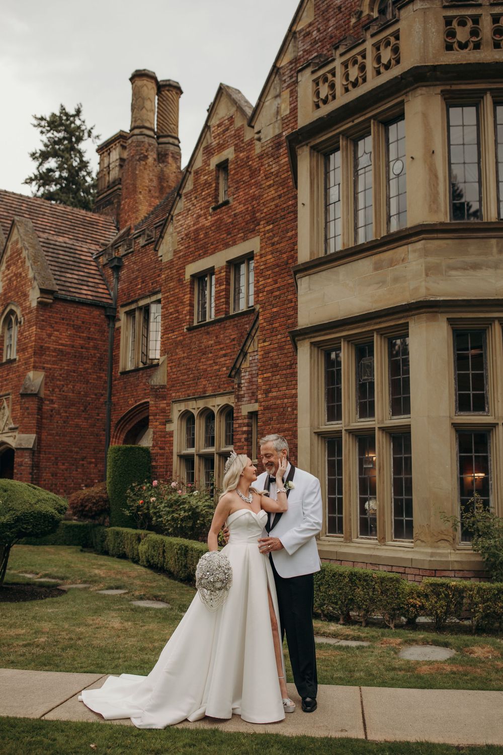 Newlyweds pose elegantly in front of a stately Victorian-style manor with red brick and stone features.