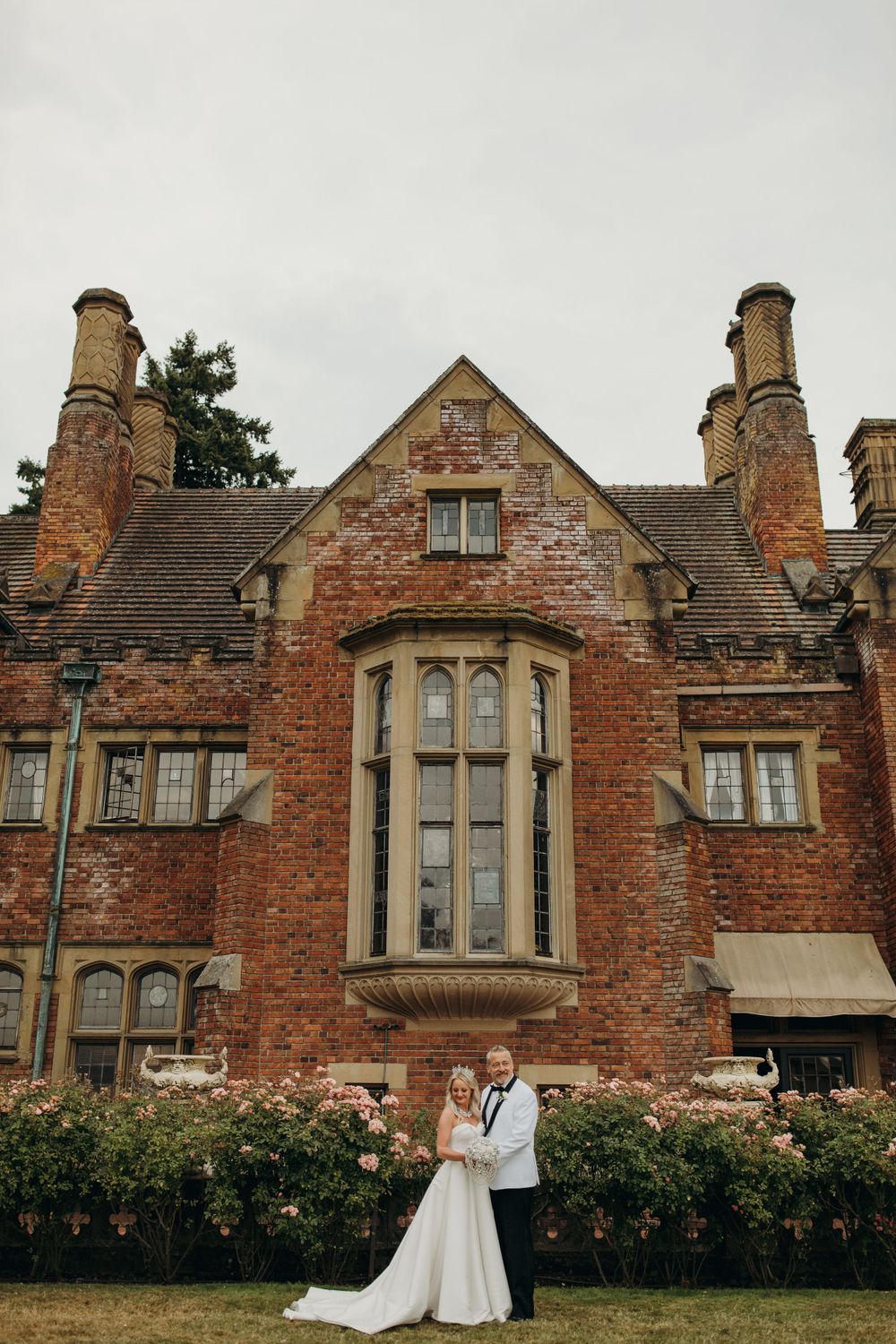Classic wedding portrait in front of historic red brick manor with tall windows and stone chimneys.