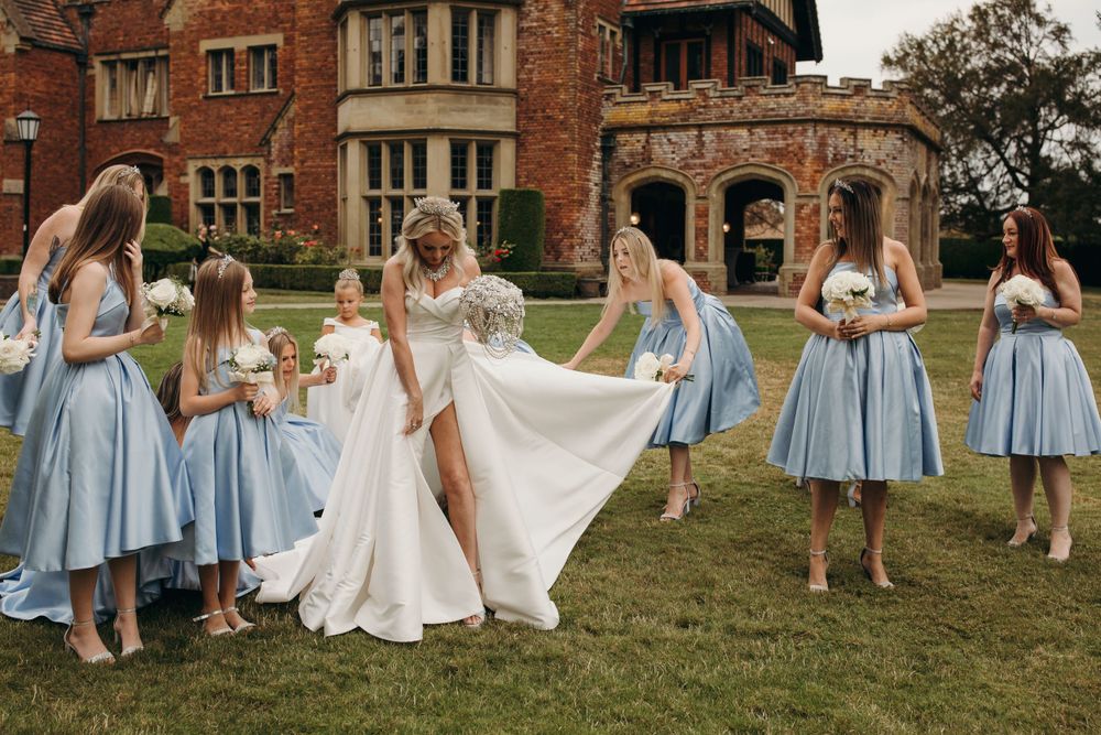 Bridesmaids in light blue dresses help arrange wedding gown on mansion lawn.