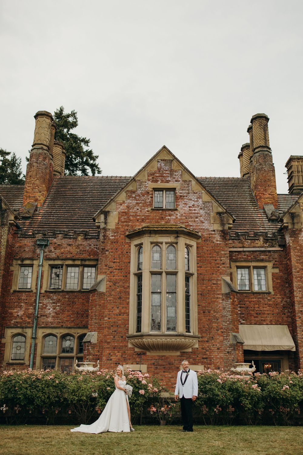 A couple poses in formal attire before a grand Gothic-style brick manor with tall stone chimneys.